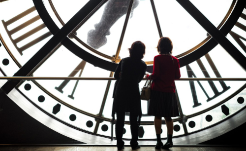 Zwei Frauen stehen auf der Innenseite einer Turmuhr und schauen durch die transparenten scheiben nach außen.