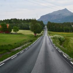 Endlos scheinende lange, gerade schmale Straße in Richtung eines Berges am Horizont. Seitlich hügelig grüne Landschaft und ein rotes gestrichenes Haus